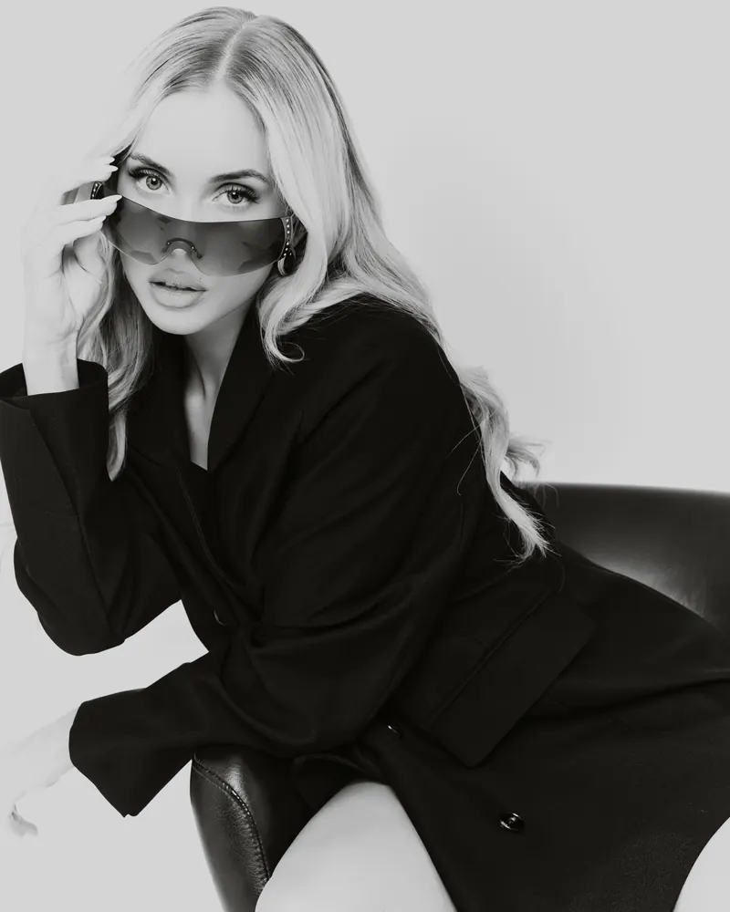 Black and white portrait of a model in a black blazer, lowering wraparound sunglasses.