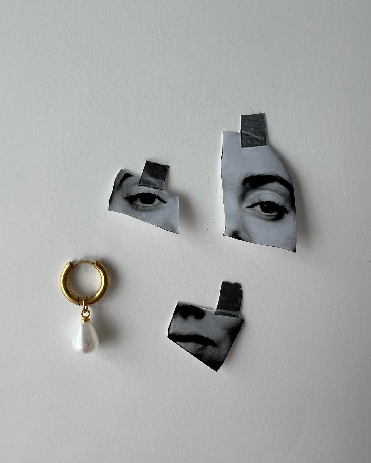 Gold hoop with a pearl drop beside cut-out paper fragments of eyes and lips.