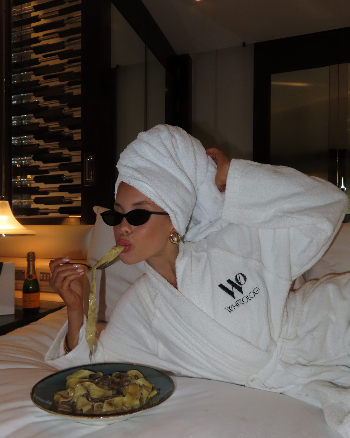 Model in a Whiteology robe and towel turban, eating pasta on a hotel bed in cat-eye sunglasses.