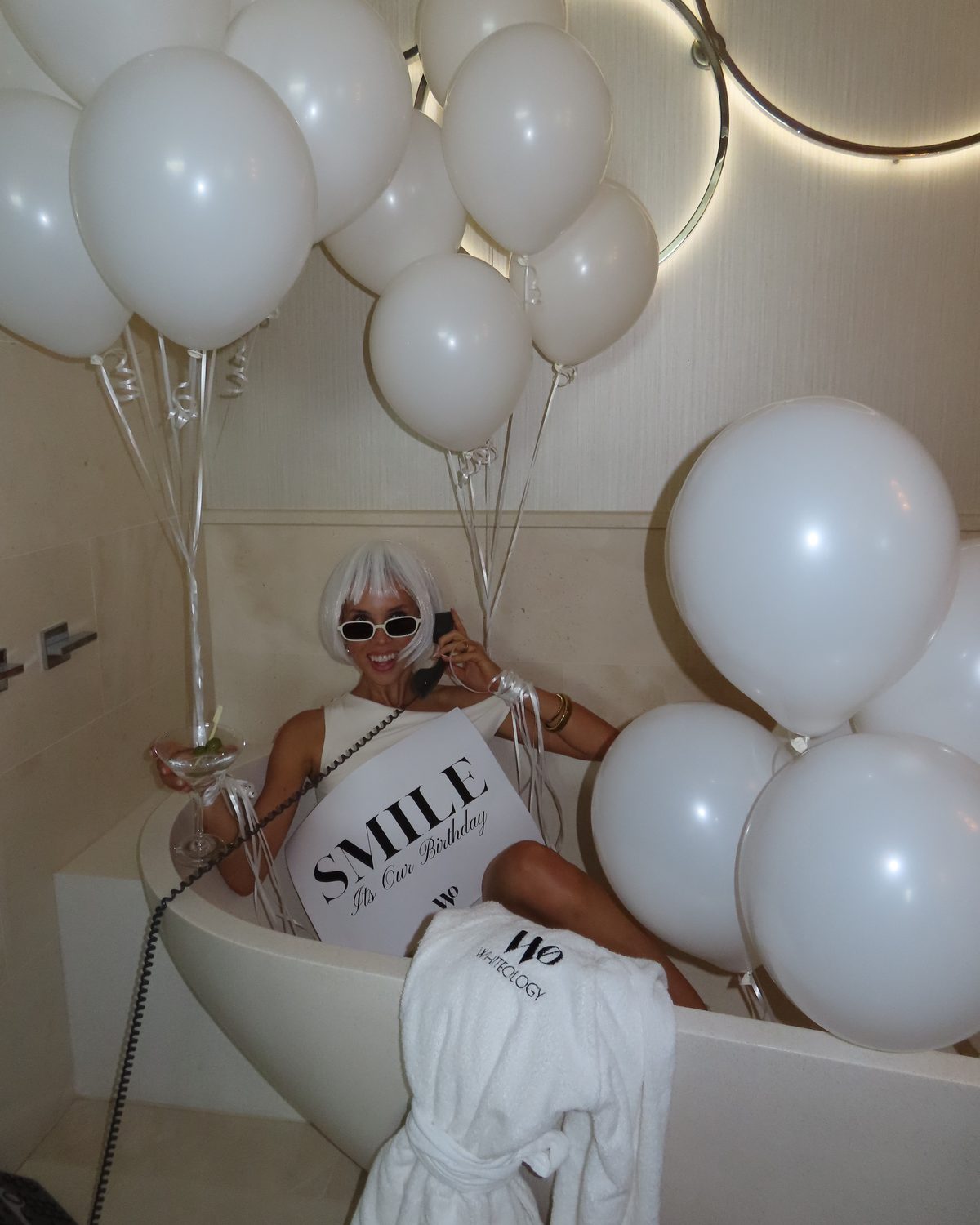 Model in a white wig and sunglasses in a bath of white balloons, holding a Smile, it's our birthday sign.